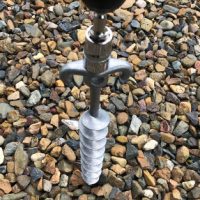 A Big Dog™ Screw In Peg, made from aluminium alloy with a left hand thread spiral stake, is partially embedded in a bed of multicolored small stones.