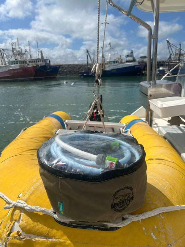 A coiled hose in a zippered storage bag rests on the bow of a yellow inflatable boat, docked at a marina with fishing boats in the background under a partly cloudy sky.