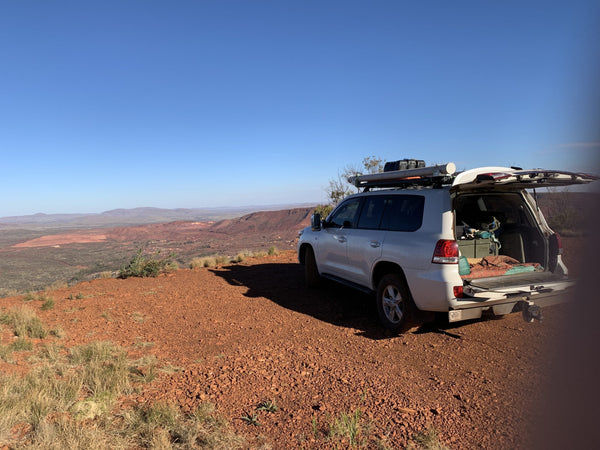 A white SUV with its trunk open is parked on a rocky, red dirt hill overlooking a vast desert landscape with distant hills under a clear blue sky.