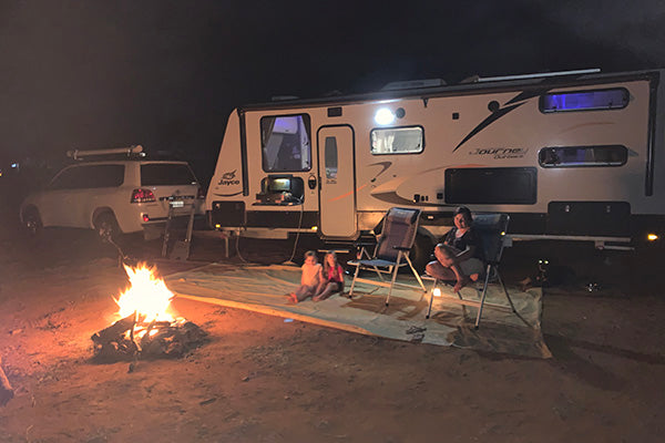 Two people relax in chairs on a mat by a campfire at night, next to a large camper trailer and a parked SUV, with the trailer lights illuminating the scene.