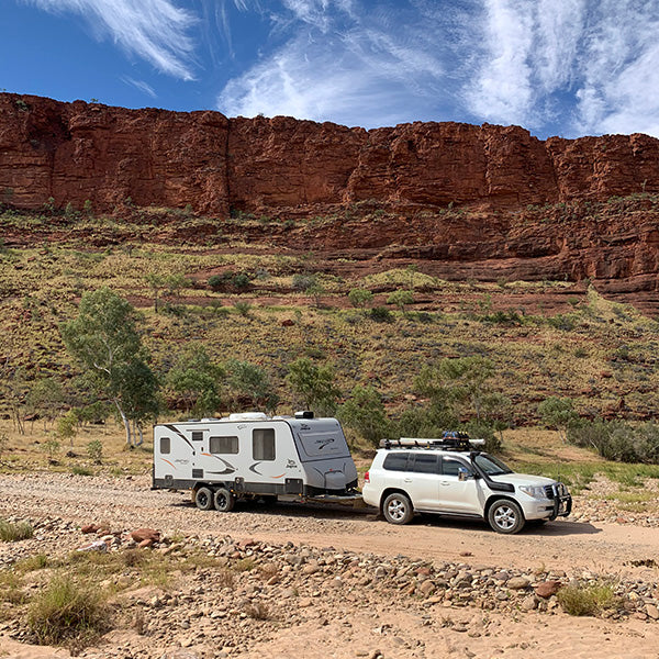A white SUV towing a gray caravan travels on a dirt road through a rocky, arid landscape with sparse vegetation and a red cliff in the background under a blue sky with wispy clouds.