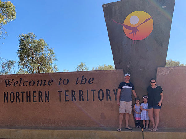 A family of four stands in front of a large “Welcome to the Northern Territory” sign under a clear blue sky, with trees and a tall monument featuring a stylized sun and figure in the background.