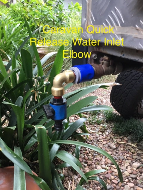 A Quick Release Water inlet elbow is attached to the side of a caravan above green plants and gravel, with yellow text labeling the device. This elbow fitting helps prevent kinking when using heavy hoses.