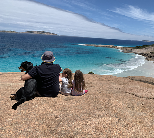 A man, two young girls, and a black dog sit on a rocky cliff overlooking a turquoise ocean and sandy beach under a partly cloudy sky.