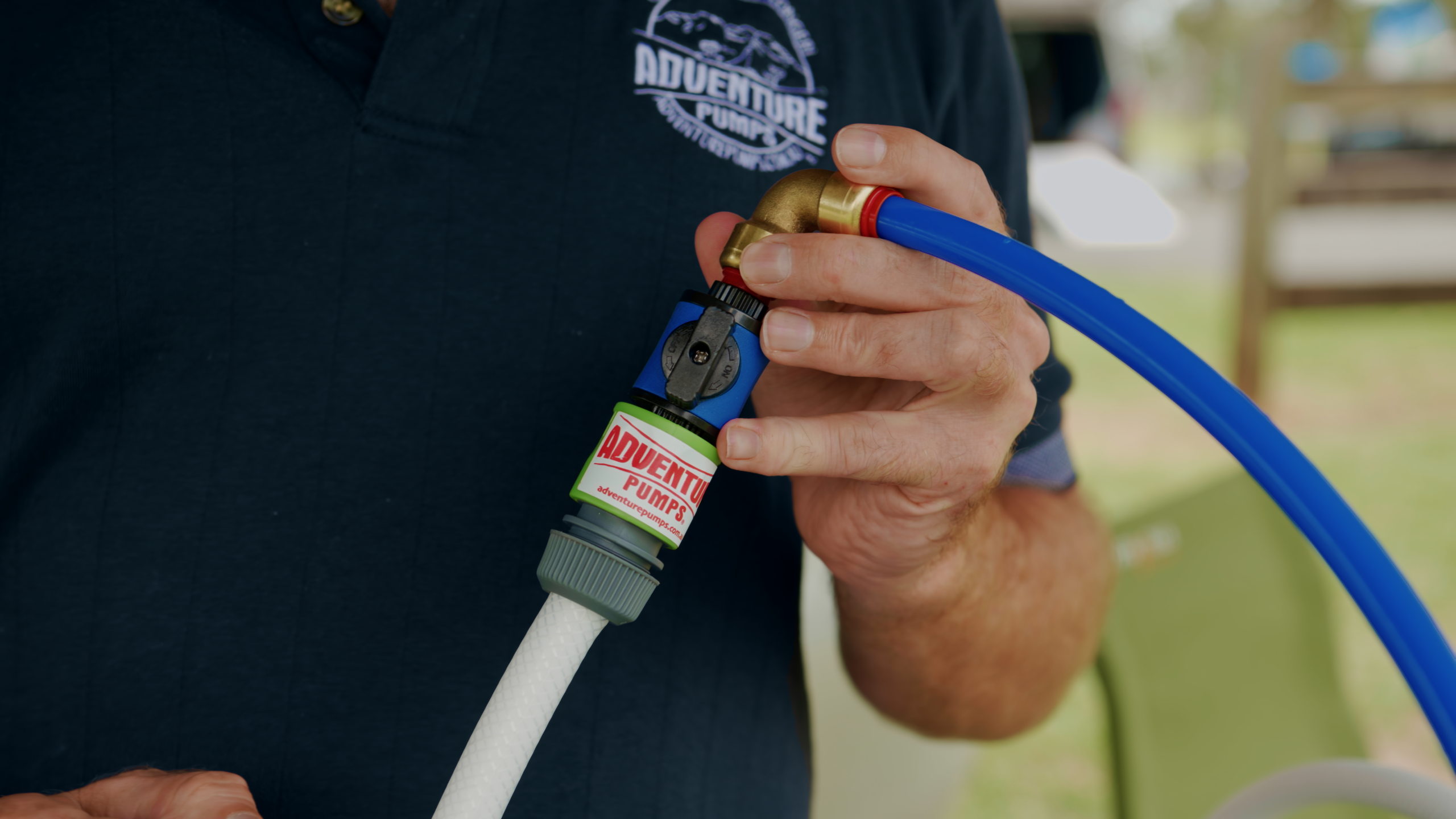 A person in a navy Adventure Pump shirt connects a blue hose with a brass fitting to a white pipe using a green and gray adapter labeled Adventure Pump.