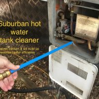 A person uses the Suburban hot water tank cleaner—a blue and brass tool—to clean a caravan’s hot water tank. Text: “Removes calcium & silt build up, even in hard to reach places. Increases heater efficiency.”.